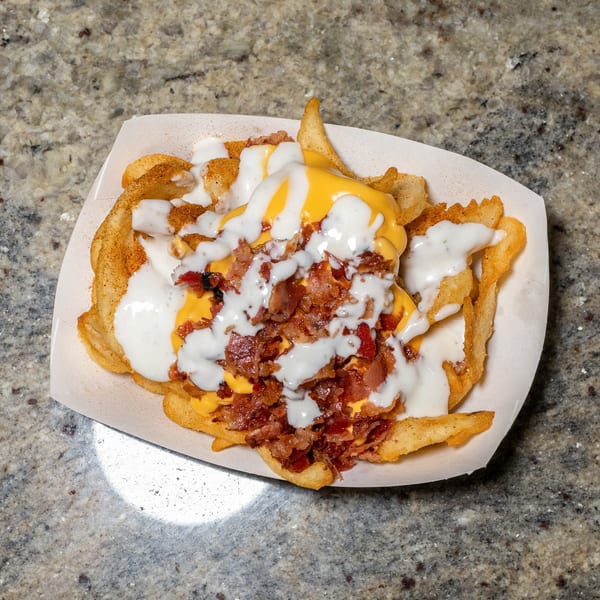 Waffle fries topped with melted cheese, bacon bits, and white sauce in a paper tray.