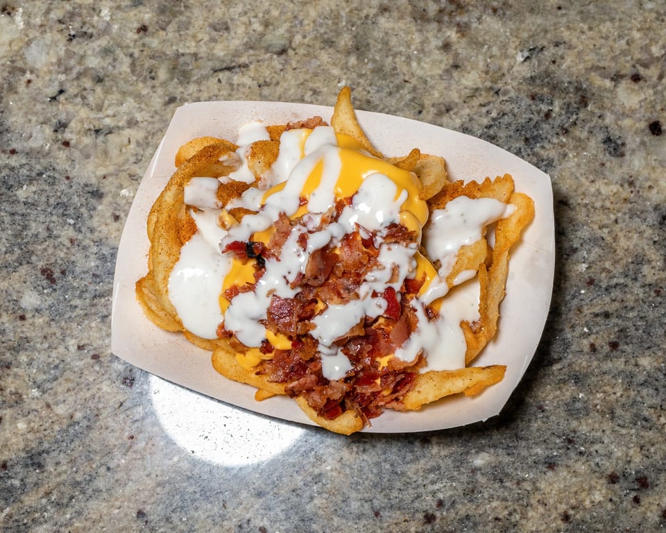 Waffle fries topped with melted cheese, bacon bits, and white sauce in a paper tray.