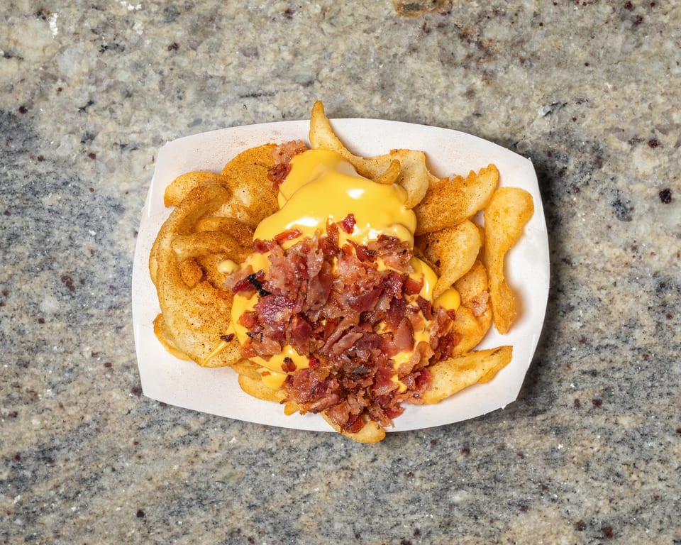Seasoned potato wedges topped with melted cheese and crispy bacon bits in a white paper tray.