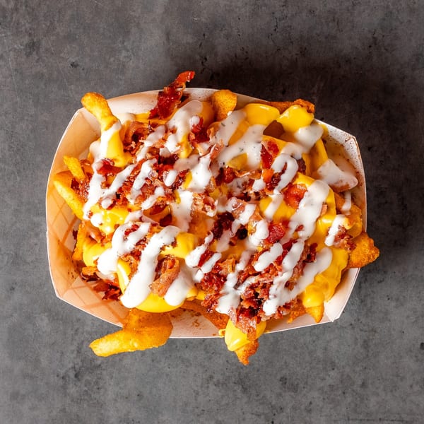 Loaded fries with melted cheese, crispy bacon bits, and a creamy white sauce in a paper tray.