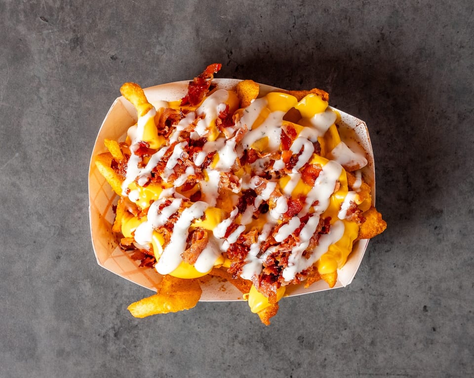 Loaded fries with melted cheese, crispy bacon bits, and a creamy white sauce in a paper tray.