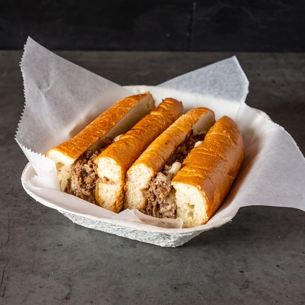 Four cheesesteak sandwiches with melted cheese and meat in a paper-lined basket.