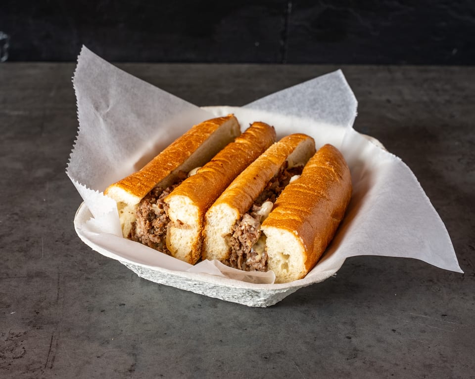 Four cheesesteak sandwiches with melted cheese and meat in a paper-lined basket.