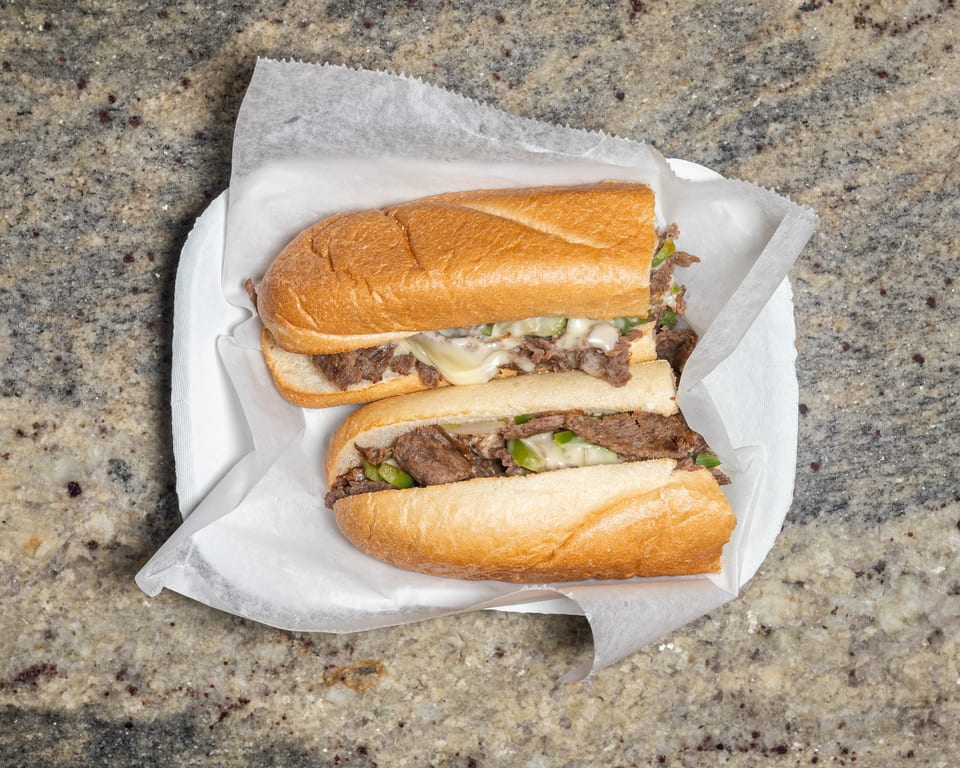 Two cheesesteak sandwiches with melted cheese, green peppers, and onions on a white plate.